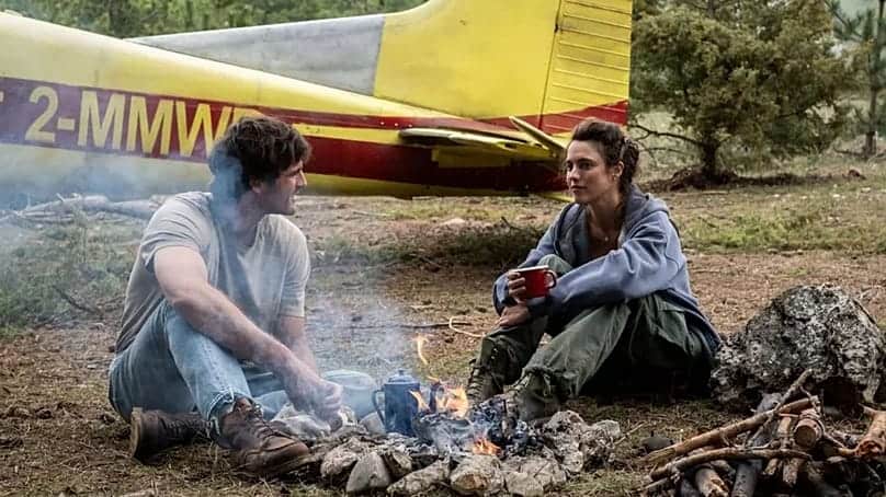 Two people sitting by a campfire with a yellow airplane in the background.